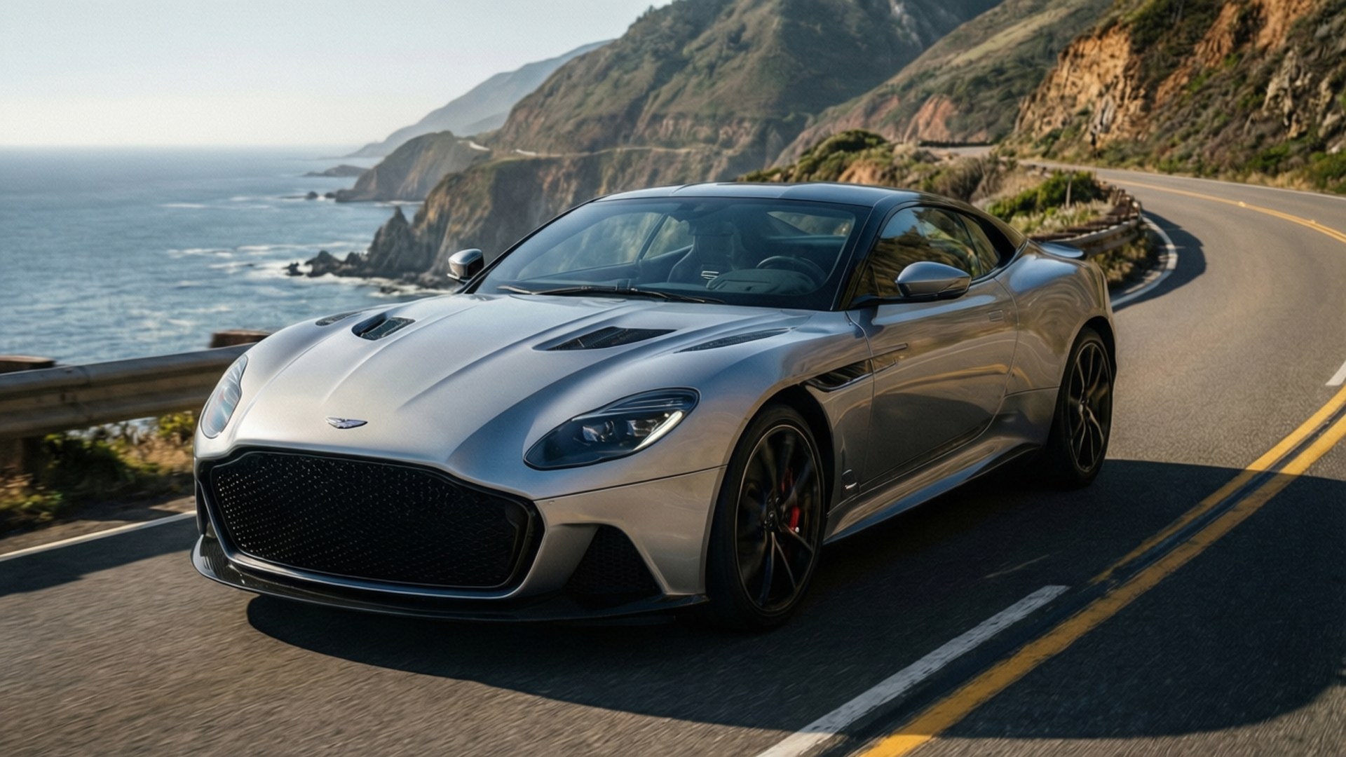 Chiếc Aston Martin DBS màu bạc uốn lượn trên con đường ven vách đá biển, ánh nắng rực rỡ.