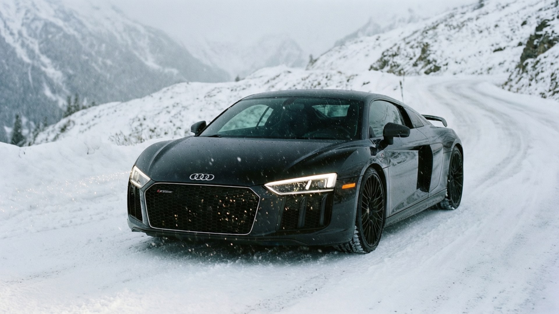 Một chiếc Audi R8 màu đen chạy trên con đường núi phủ đầy tuyết với tuyết rơi.